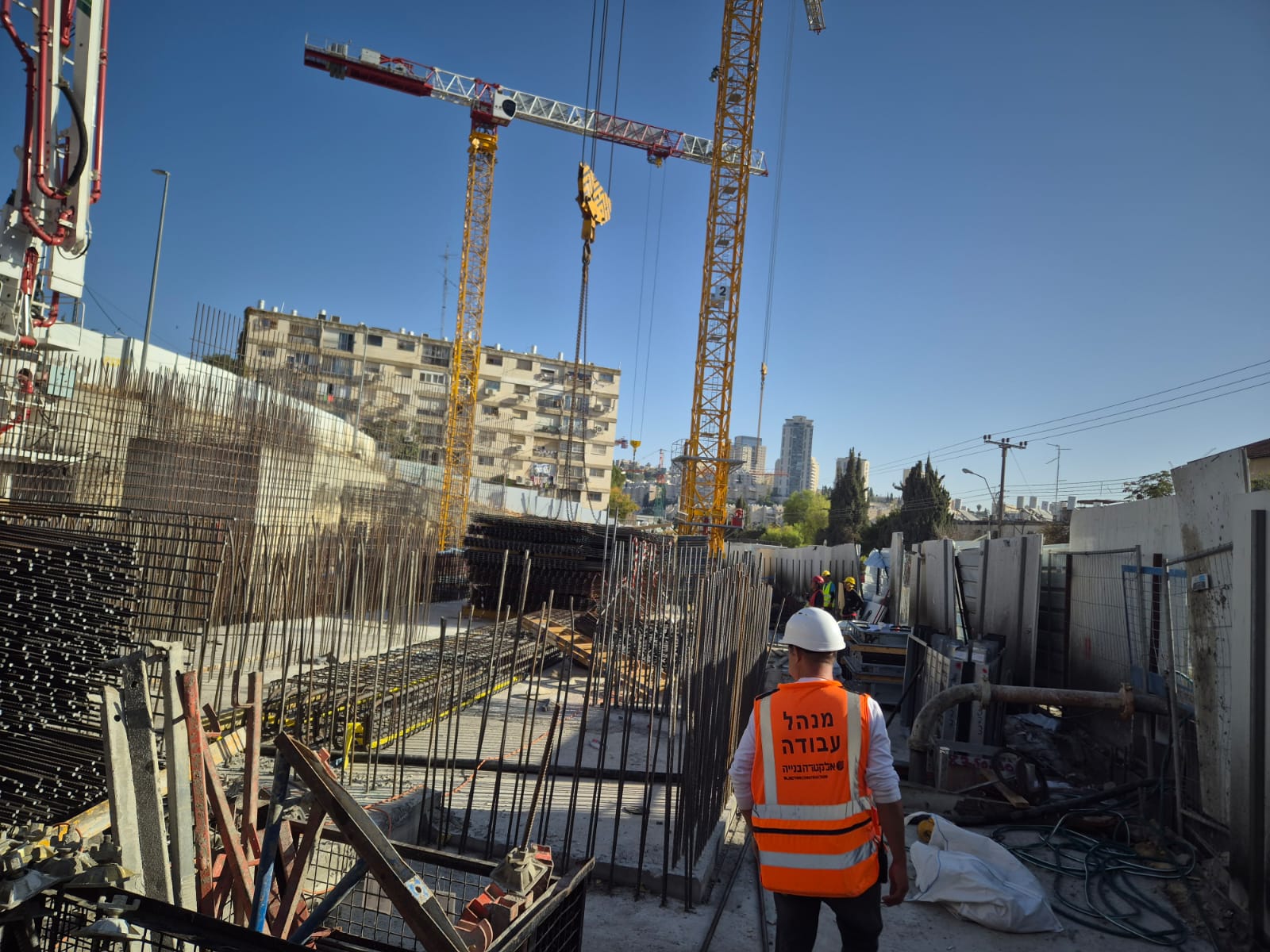 עובד באתר בנייה עם קסדה ווסט זוהר, בניין מתהווה עם מנופים ברקע.