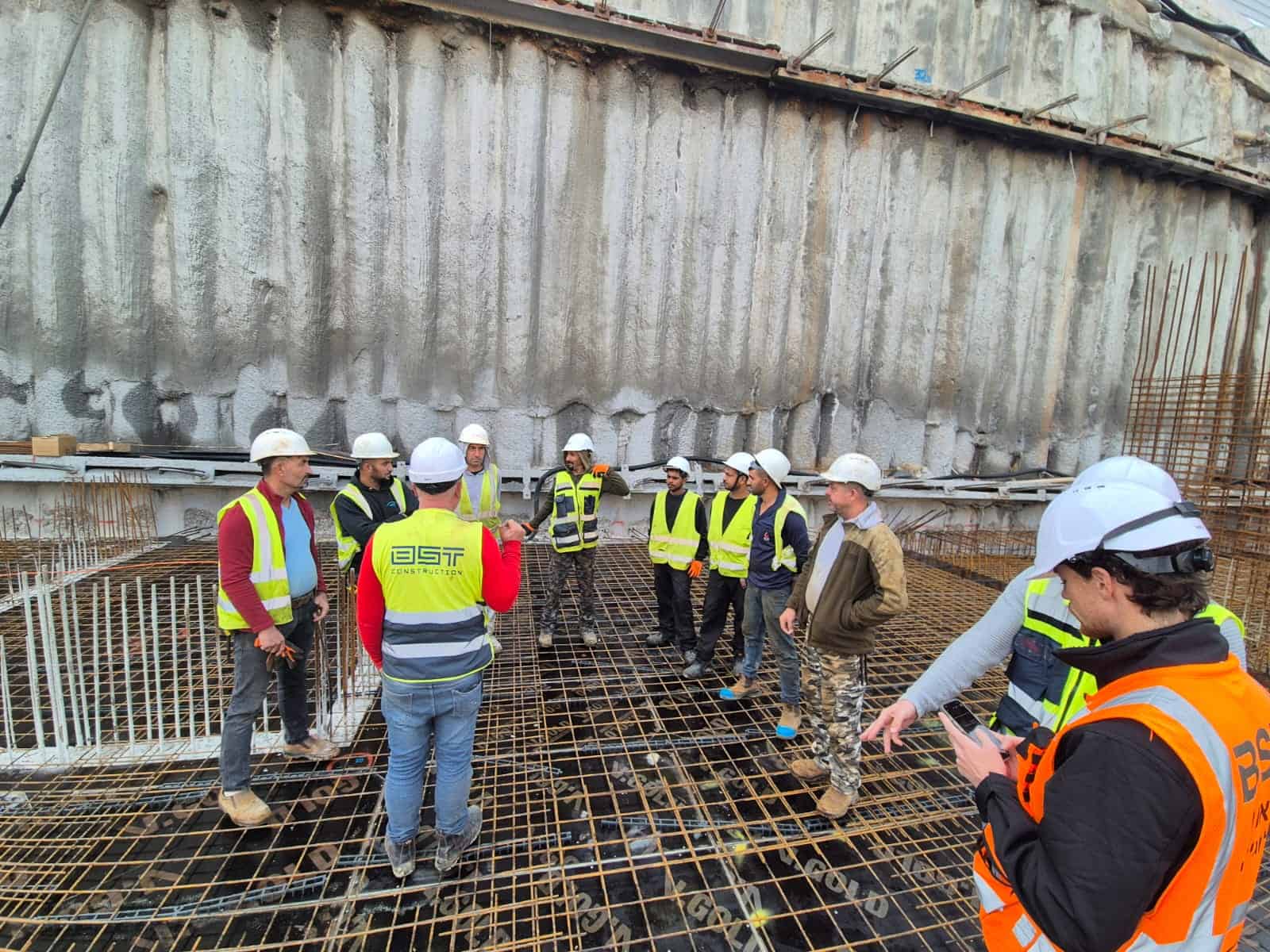 קבוצת עובדים באתר בנייה עם ציוד מגן ומבנה תעשייתי ברקע.